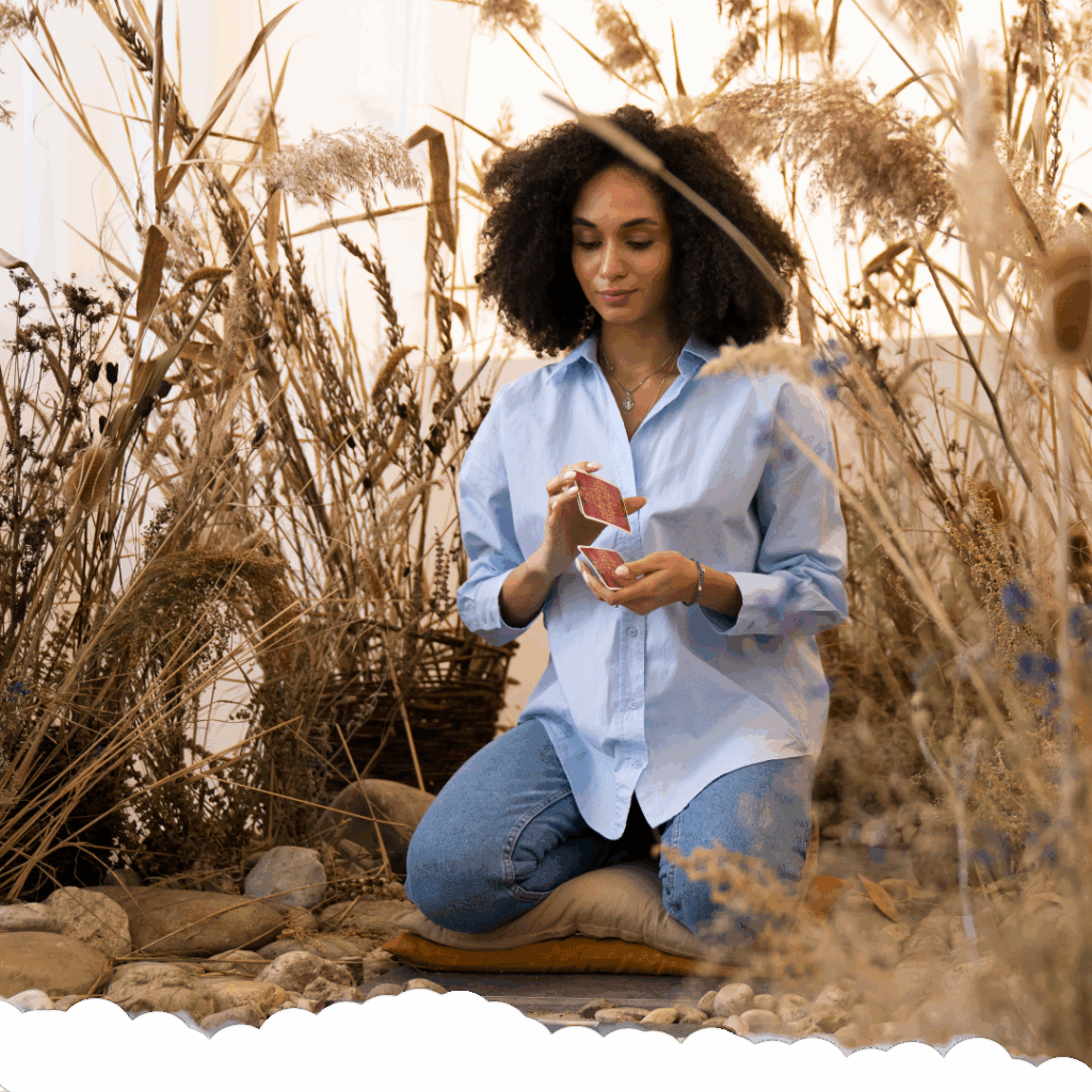 A woman with curly hair sits cross-legged on a cushion among dried wild grasses, holding and shuffling oracle or tarot cards, creating a serene and reflective atmosphere.